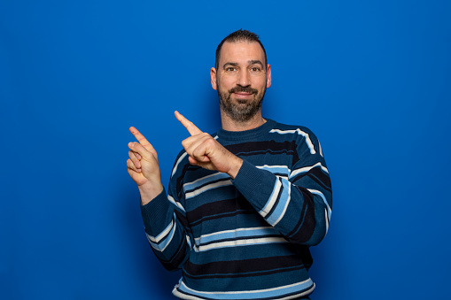Excited young bearded man in striped pullover posing isolated on blue background studio portrait. People candid emotions lifestyle concept. Mock up copy space. Point index fingers to the side.