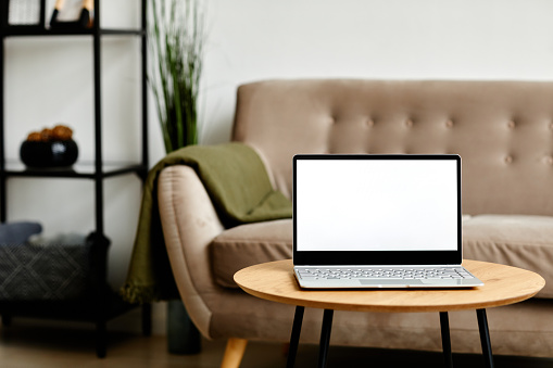 Background image of open laptop with blank white screen on coffee table in living room interior, copy space