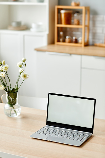 Vertical background image of white laptop screen on wooden table in kitchen decorated with fresh flowers in Spring, copy space