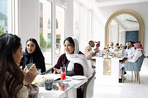 Focus on foreground group of Middle Eastern friends in traditional attire sitting at window table enjoying juice and light lunch.
