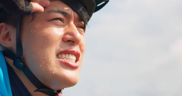 close up of asian young male cyclist wearing helmet feel tired and wipe his sweat - sunshine on his sweating face
