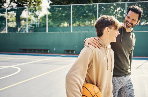 father and son walking after playing a game of basketball. young man and teenage boy having fun, talking and chatting while staying fit, active - zoon stockfoto's en -beelden