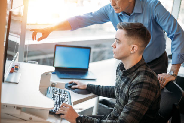 colega que asesora a un asociado más joven en la oficina de negocios mientras señala los datos en el monitor de la computadora - aprendiz fotografías e imágenes de stock