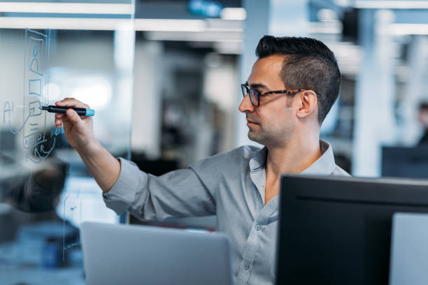 istock Mature man writing ideas on transparent glass sketch board in modern office with computers 1395838927