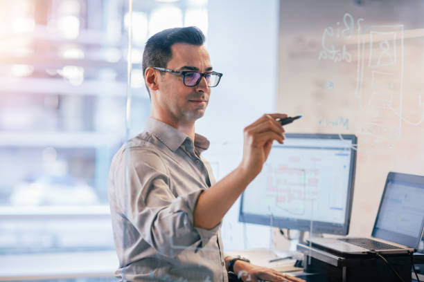 istock Mature man writing ideas on transparent glass sketch board in modern office with computers 1395839001