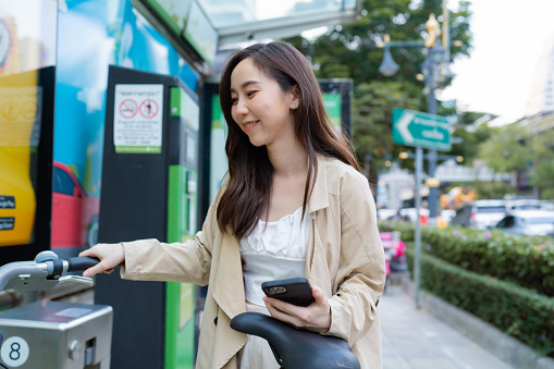 Young Asian woman using smart phone contactless payment at public bicycle service station in the city.