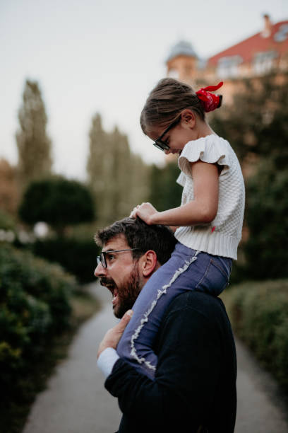 Being goofy is her style Spontaneous and funny photo of a young, bearded, father playing with his little daughter while she pulls his hair, sitting on his shoulders and laughing, he portrays pain on his face. child laughing hysterically stock pictures, royalty-free photos & images