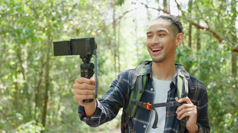 Asian handsome male backpacker vlogger traveling in the forest alone. Attractive man traveler use mobile phone record video vlogging, walking in nature wood with happiness during holiday vacation trip