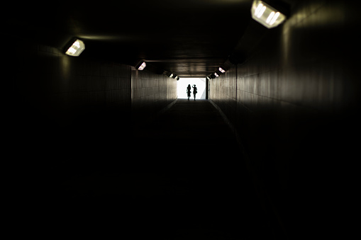 Two silhouettes at end of tunnel. Light in distance. Figures of small women in contrasting light. Lamps in long underpass.