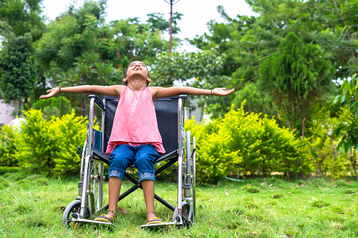 concept of freedom, individuality and courage showing by Happy girl kid feeling nature by stretching arms while using wheelchair at hospital park.