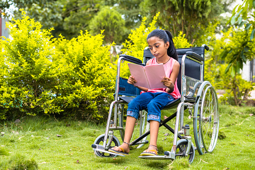 Teenager girl kid with disability reading book while using wheelchair at garden - concept of education, development and learning