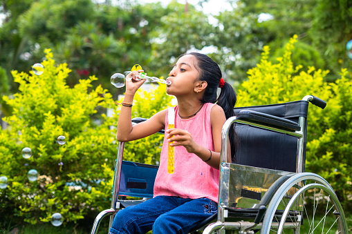 cheerful girl kid playing by blowing water bubbles while using wheelchair at park - concept of playful activities, happiness and inspiration.