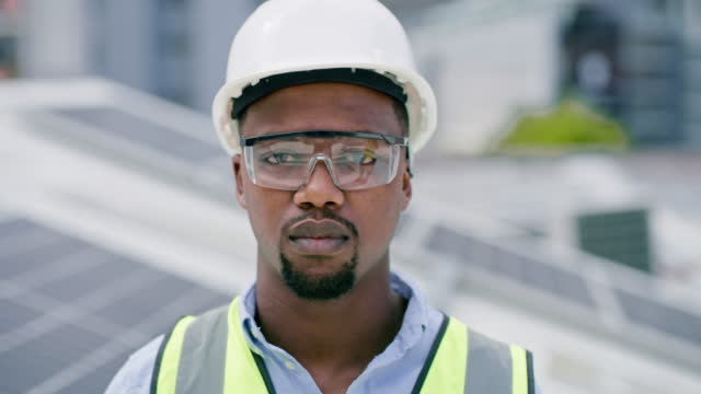 A young african american man wearing goggles and a hardhat while installing a solar panel. Portrait of a confident male employee standing on a building at a construction site