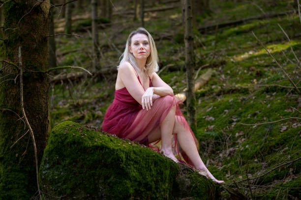 Illuminated side view portrait of beautiful confident blond woman wearing magenta gown sitting on moss covered rock at forest