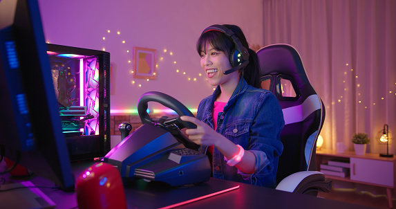 young asian pretty pro gamer wearing headset is playing racing game with friends online at home