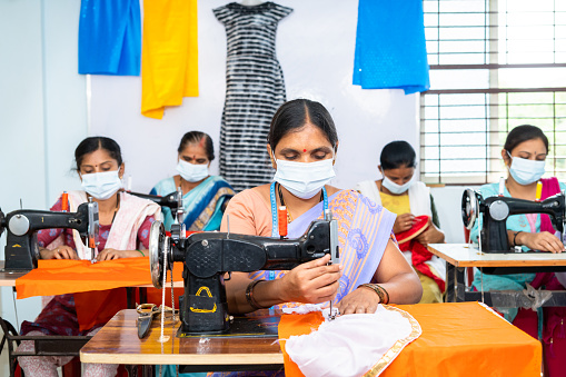 group of garments workers in medical face mask buys working at garments on sewing to protect from coronavirus or covid-19 outbreak - concept of social distance, back to work and safety.