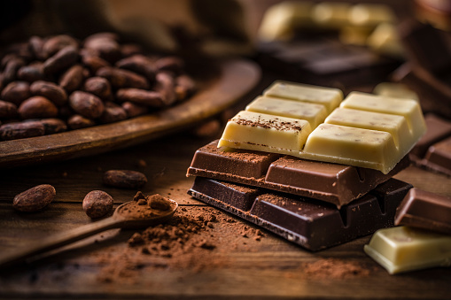 Front view of  a stack of three types of chocolate bars surrounded by cocoa powder and cocoa beans on a rustic wooden table. The composition includes a dark chocolate bar, a milk chocolate bar and a white chocolate bar.