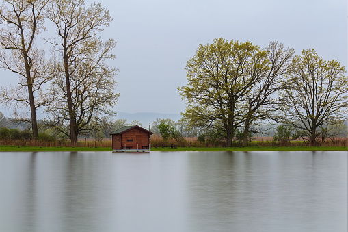Wooden cabin, building on pond shore between trees in rain moody weather. Long exposure, Czech landscape