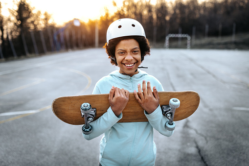 Portrait of cheerful carefree girl wearing sports helmet holding skateboard in park at sunset