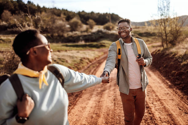 Male explorer holding girlfriend's hands hiking on dirt road Happy young male explorer with backpack holding girlfriend's hands while hiking on dirt road in summer hiking bandana stock pictures, royalty-free photos & images