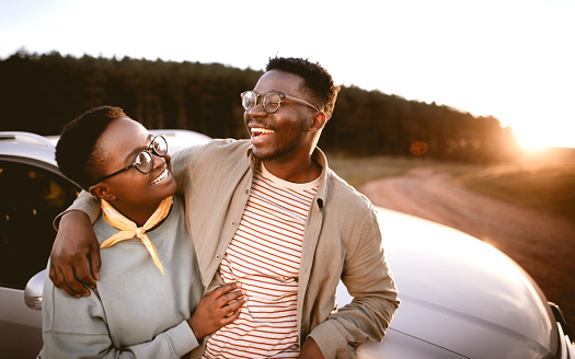 Happy young man with hand on girlfriend's shoulder looking away while standing by car against clear sky during sunset