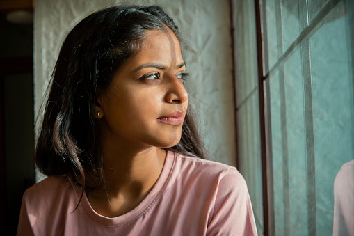 Indoor close-up portrait of beauty, Asian, Indian serene young woman sitting near the window and contemplating while looking out through the window.