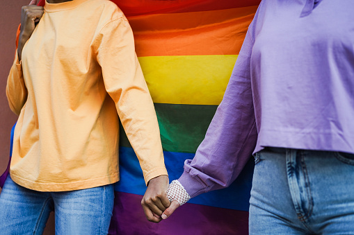 Happy women gay couple holding hands with rainbow flag on background - Focus on hands