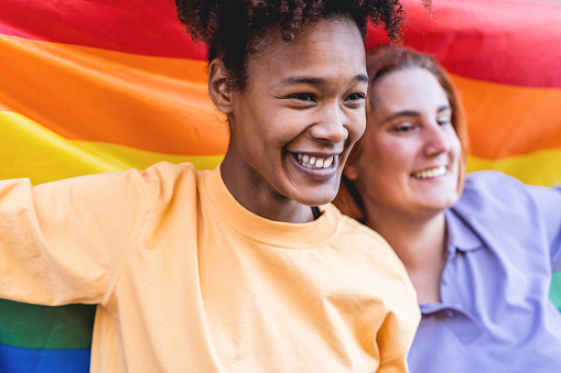 Happy women gay couple holding rainbow flag outdoor - Focus on african girl face