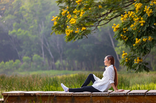 Asian woman sitting on the edge of dock with peaceful natural park during summer with yellow flower blossom for serene and relaxation outdoor recreation concept