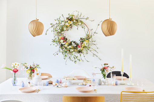 A modern table decoration as an Easter table with a floral Easter wreath hanging on the wall in a bright, large room. Photographed in high resolution