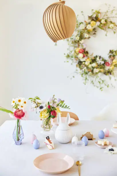 A modern table decoration as an Easter table with a floral Easter wreath hanging on the wall in a bright, large room. Photographed in high resolution