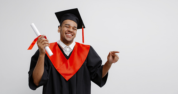 A Black student in graduate gown and square cap who is happy to finish his studies, shows his the long-awaited diploma