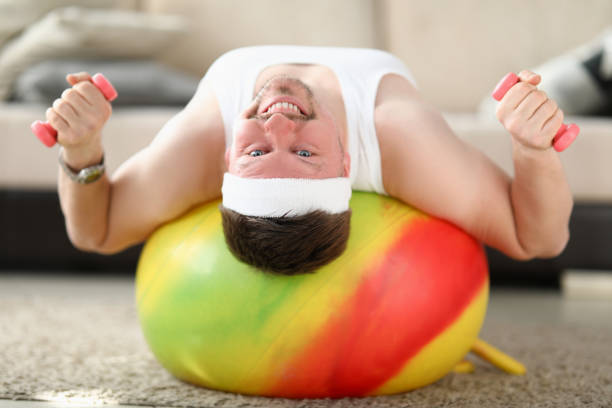 Portrait of middle-aged man training at home on fitness ball. Person lifting dumbbells. Living room interior. Goal for strong and fit body. Sport and active lifestyle concept