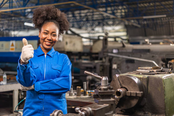 工場industrial.peopleで保護制服操作機を着たアフリカ系アメリカ人の若い女性労働者は、産業で働いています。職場でカメラを見つめる女性エンジニアの肖像画。 - heavy plant ストックフォトと画像