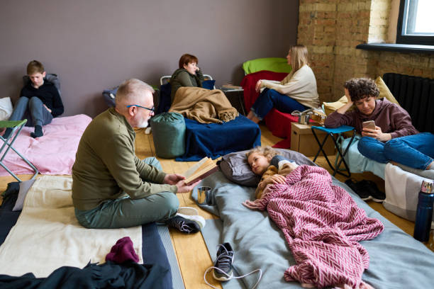 Mature man with open book reading fairy tale to his grandson lying in bed Mature man with open book reading fairy tale to his grandson lying in bed while resting in temporary shelter with other refugees sleeping grandpa stock pictures, royalty-free photos & images