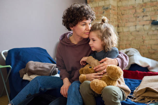 Young woman giving hug to her cute little son with brown soft teddybear Young woman giving hug to her cute little son with brown soft teddybear while both sitting on sleeping place prepared for refugees temporary stock pictures, royalty-free photos & images