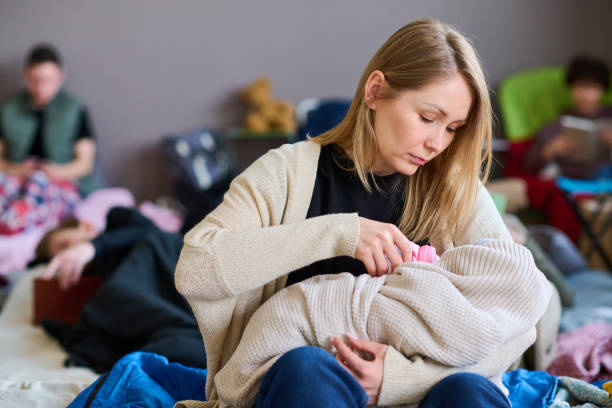 Young woman with long blond hair feeding her baby wrapped into warm plaid Young woman with long blond hair feeding her baby wrapped into warm plaid with milk while sitting in front of camera against refugees emergency shelter stock pictures, royalty-free photos & images