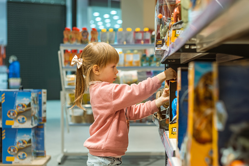 Little caucasian girl choosing a new toy in the big baby store. Big shelfs full of toys