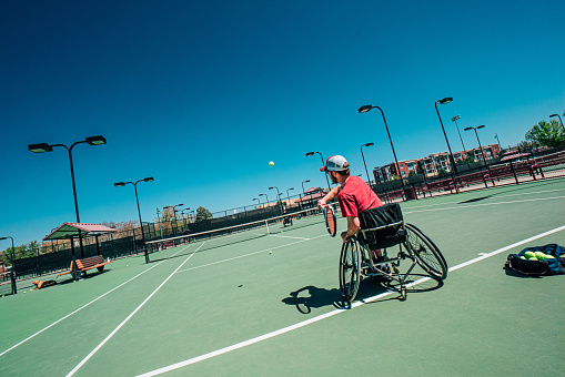 Handsome Young Man with Myelomeningocele Spina Bifida Sitting in a Wheelchair Practicing and Playing Tennis for Adaptive Exercise Outdoors in the Summer. Overcoming the Stigma and Challenges of Disability can be Fun on the Tennis Court!