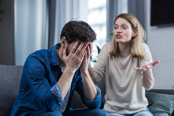 conflitto familiare, litigio, incomprensione. la donna grida al marito, disperato, piangendo. richiede spiegazioni. l'uomo ascolta, coprendosi il viso con le mani. seduto sul divano di casa. - relationship difficulties couple anger communication breakdown foto e immagini stock