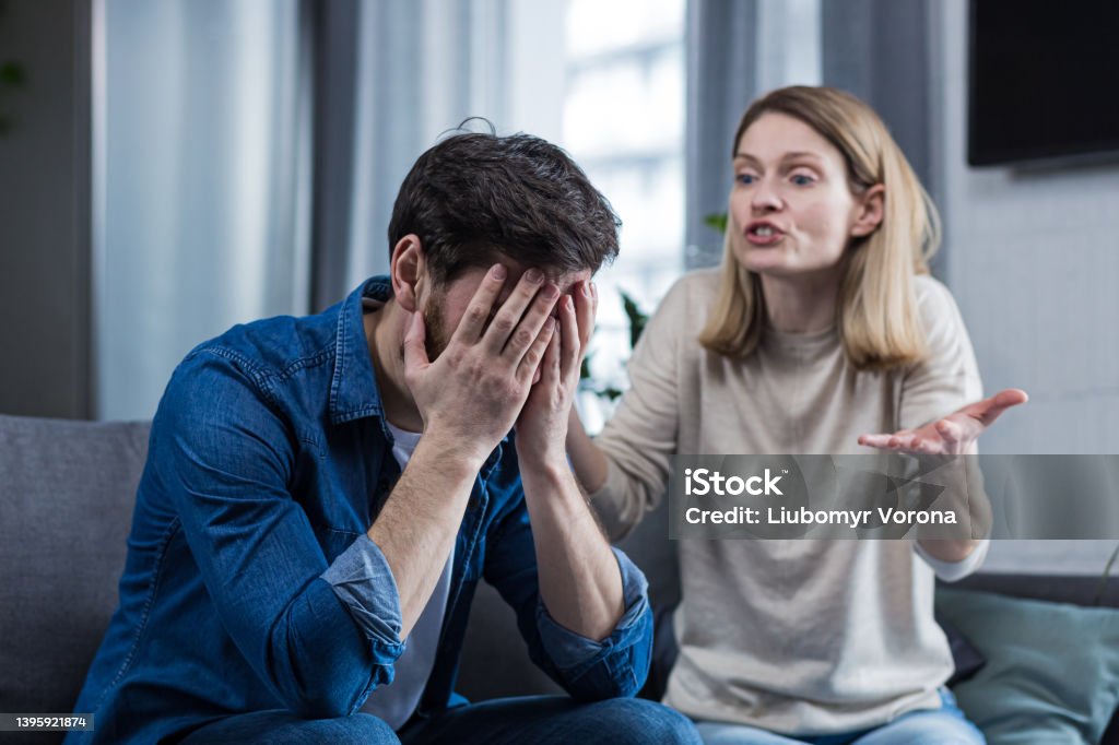 Family conflict, quarrel, misunderstanding. The woman shouts at her husband, in despair, crying. requires explanation. The man listens, covering his face with his hands. Sitting on the couch at home. Couple - Relationship Stock Photo