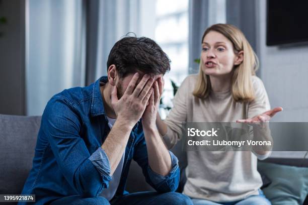 Family Conflict Quarrel Misunderstanding The Woman Shouts At Her Husband In Despair Crying Requires Explanation The Man Listens Covering His Face With His Hands Sitting On The Couch At Home Stock Photo - Download Image Now