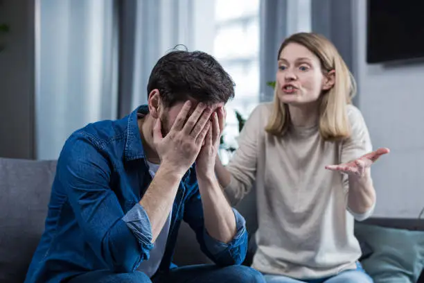 Photo of Family conflict, quarrel, misunderstanding. The woman shouts at her husband, in despair, crying. requires explanation. The man listens, covering his face with his hands. Sitting on the couch at home.