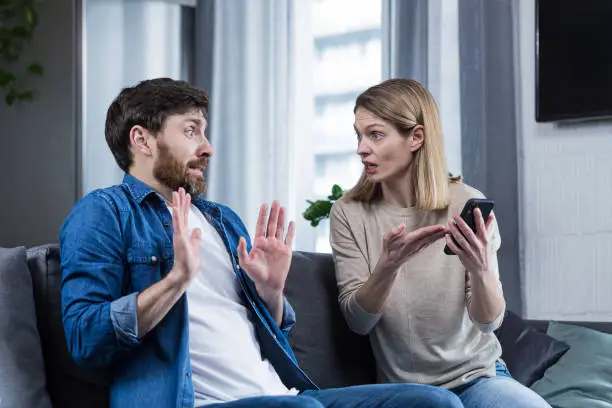 Photo of Family conflict. The woman suspects her husband of treason, looks at his phone, demands explanations. sitting at home on the couch.