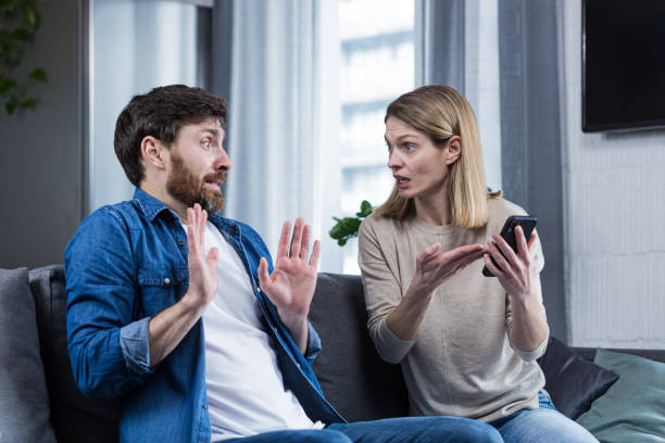 family conflict. the woman suspects her husband of treason, looks at his phone, demands explanations. sitting at home on the couch. - infidelidade imagens e fotografias de stock