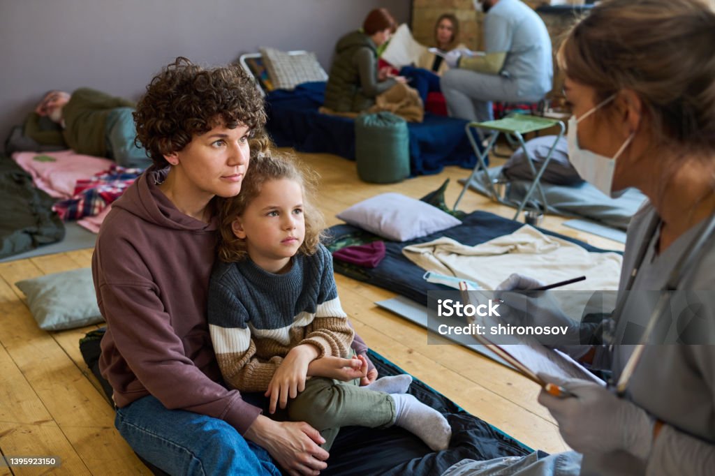 Young female refugee with son looking at clinician making notes in document Young female refugee with little son looking at clinician in uniform and mask making notes in medical document and consulting them Homelessness Stock Photo