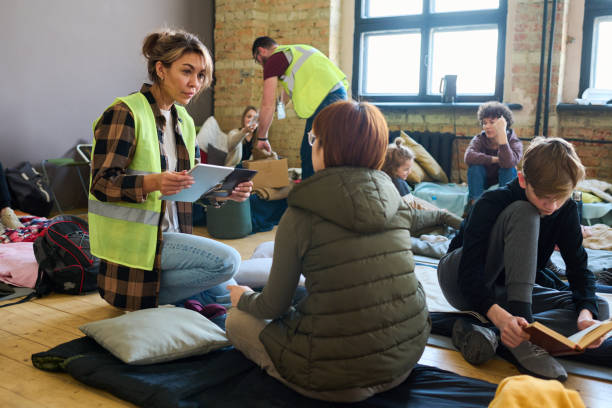 Female volunteer with mobile gadgets communicating to one of refugees Female volunteer with mobile gadgets communicating to one of refugees while sitting on squats in front of her in shelter for migrants deportation stock pictures, royalty-free photos & images