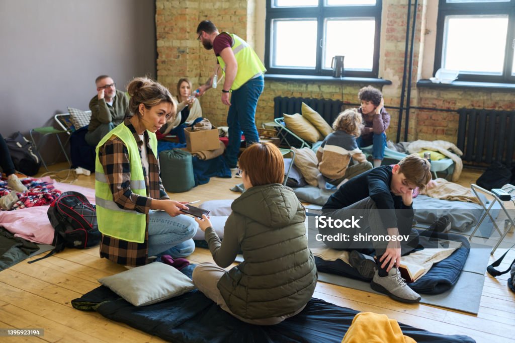 Young female volunteer passing smartphone to one of refugees sitting on mattress Young female volunteer passing smartphone to one of refugees sitting on mattress on the floor of spacious room serving as camp Refugee Stock Photo