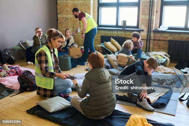 Young Female Volunteer Passing Smartphone To One Of Refugees Sitting On Mattress Stock Photo - Download Image Now
