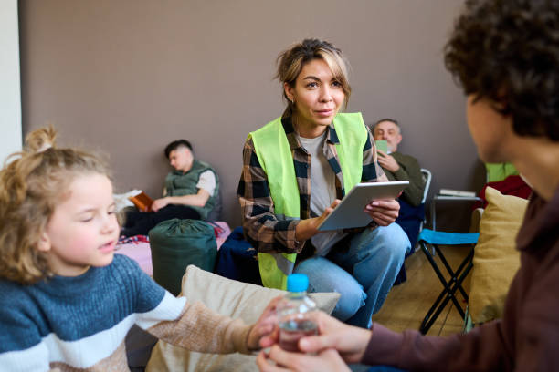 Young female volunteer with tablet sitting on squats in front of woman with son Young female volunteer with tablet sitting on squats in front of woman with son and entering their names and other personal data emergency shelter photos stock pictures, royalty-free photos & images
